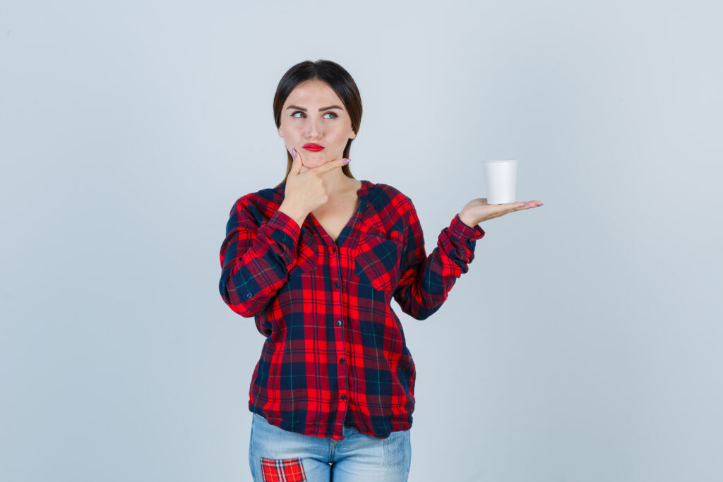 Portrait of young beautiful female holding plastic glass, keeping hand under chin in casual shirt, jeans and looking pensive front view. How Long Can Milk Sit Out.